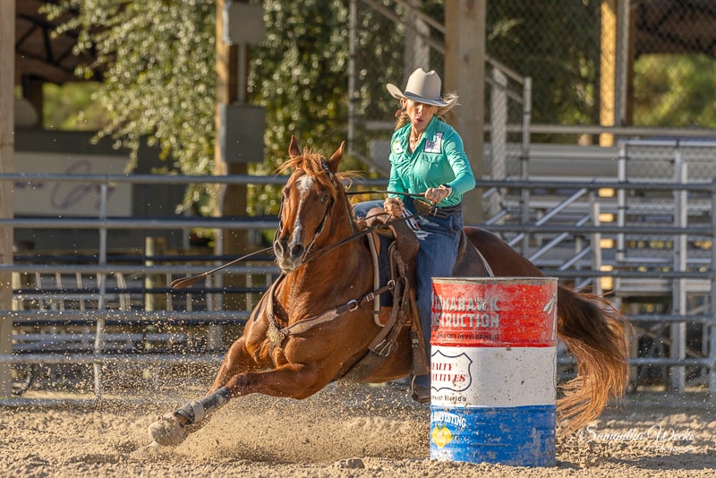 Cowgirl in teal shirt on chestnut horse kicking up dirt at Tomahawk arena daytime