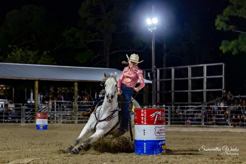 Cowgirl in pink shirt and black helmet on grey horse rounding 65th Cracker Day Rodeo barrel at night