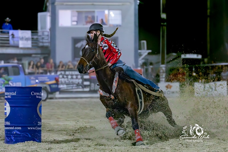 Rider in red plaid on dark horse kicking up dirt around barrel
