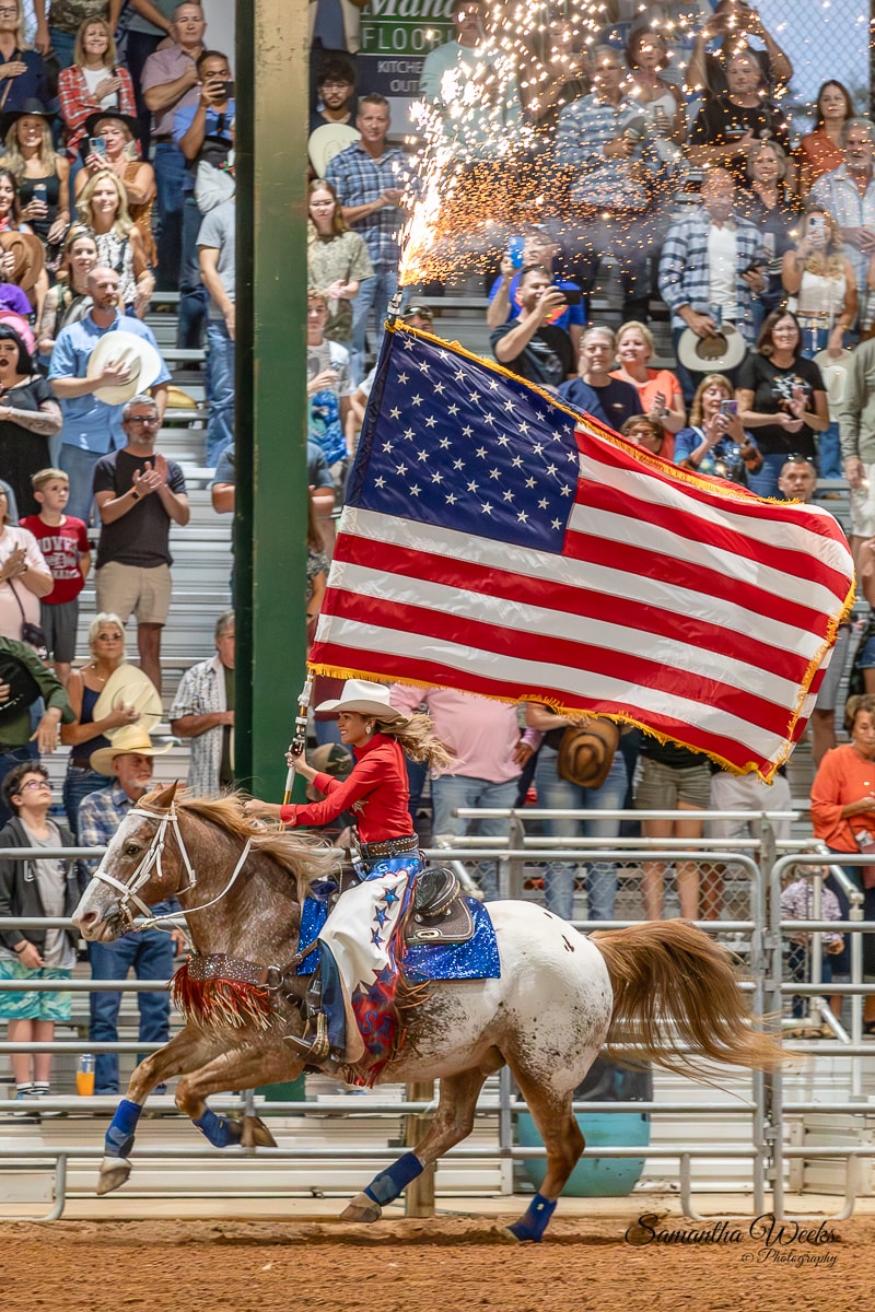 Cowgirl on appaloosa horse carrying American flag with pyrotechnic sparks before packed crowd