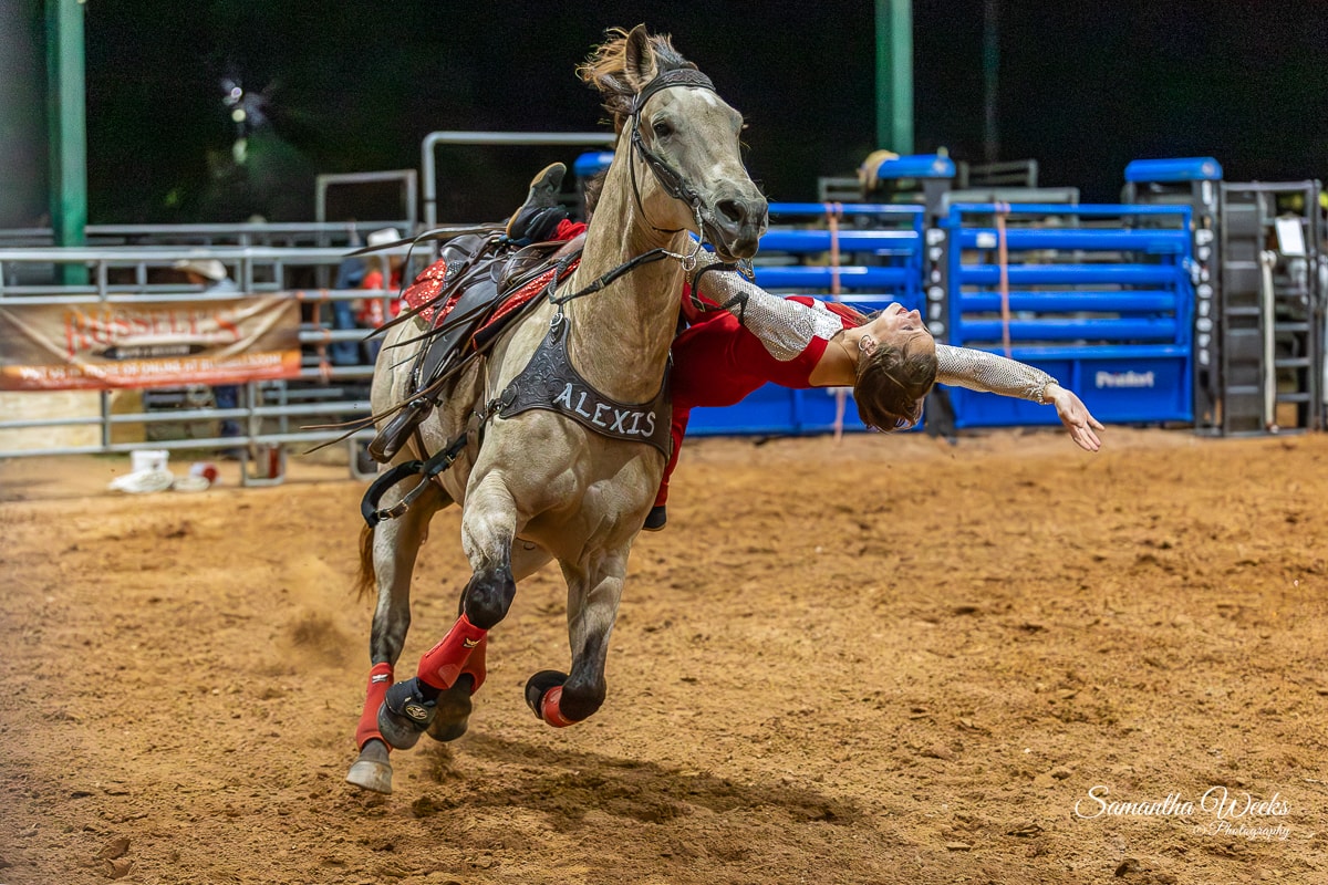 Trick rider named Alexis leaning back off grey horse at full gallop in arena at night