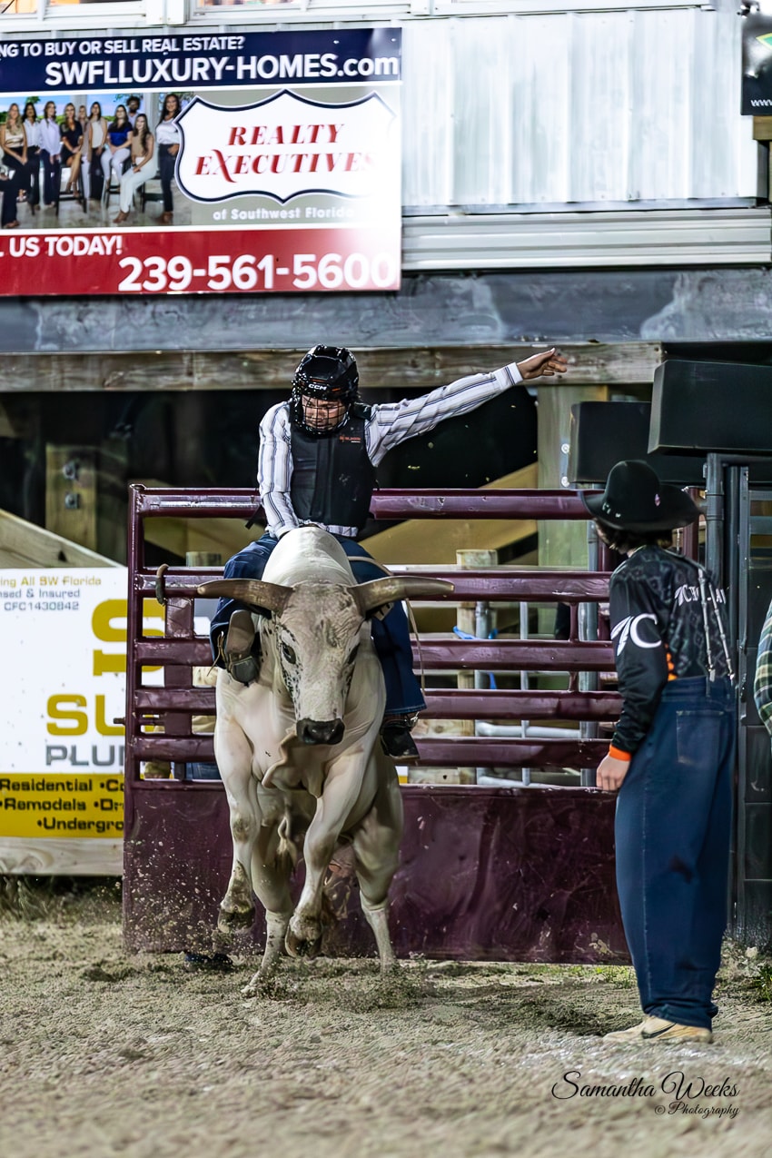 Bull rider in black helmet on white longhorn bull bursting from gate at night rodeo